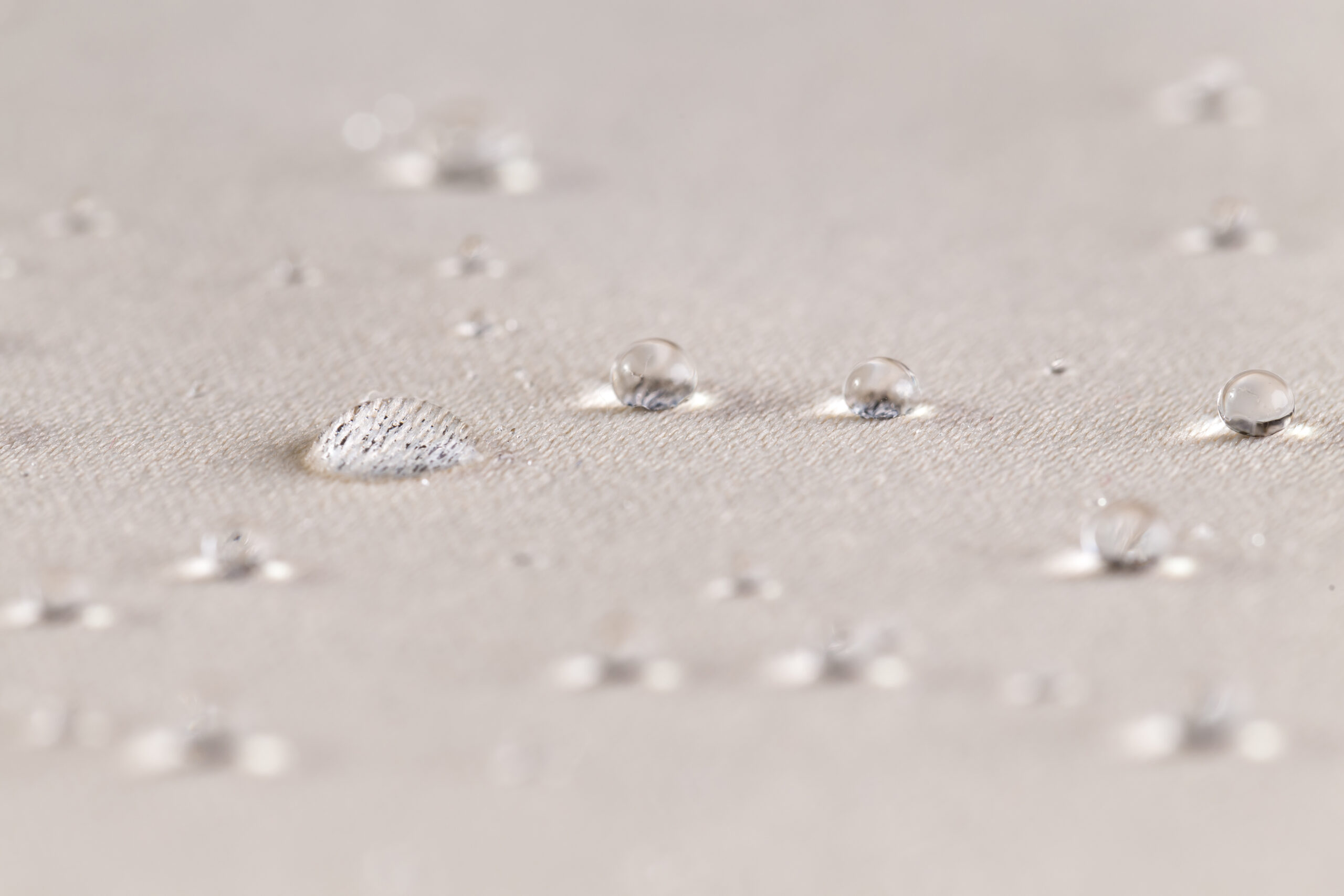 Pequeñas gotas de agua se agrupan sobre una superficie beige texturizada, mientras una gota mayor resbala y refleja la luz con nitidez.