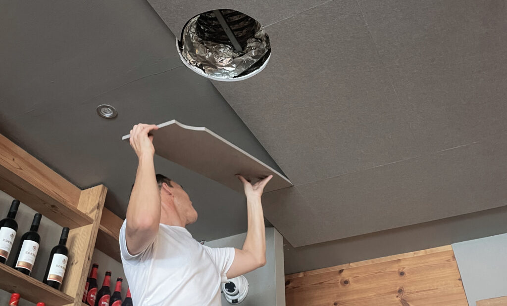 Un técnico instala paneles de aislamiento acústico en un techo interior mientras trabaja junto a una apertura de ventilación en un local.