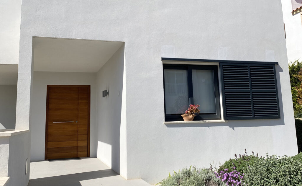 Fachada blanca con puerta de madera, ventana negra con persiana y entrada cubierta en una vivienda moderna con acabado de aislamiento térmico.