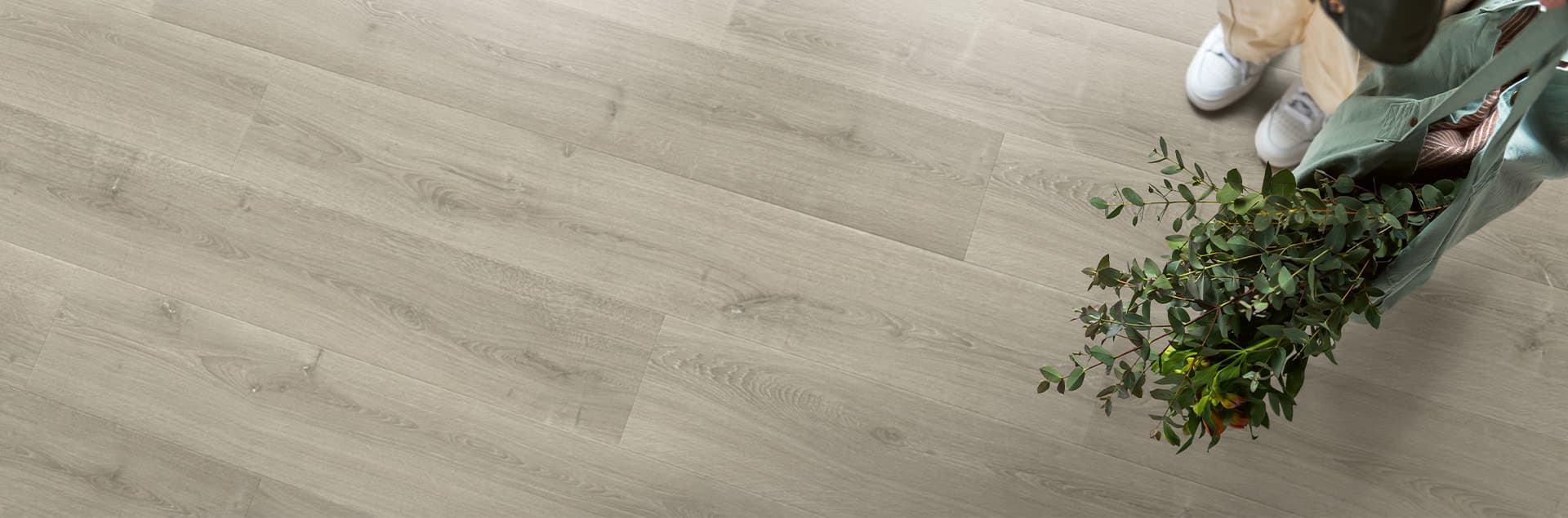 Un suelo laminado gris claro de Quick-Step se ve junto a unas piernas con zapatillas blancas y un ramo verde sobre la tarima en primer plano.