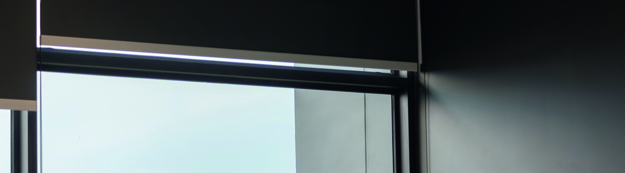 Una ventana con persiana enrollable negra deja pasar luz suave sobre un espacio interior de tonos grises y negros, con acabado moderno.