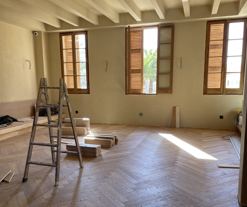 Suelo de parquet en espiga claro y estancias en reforma con ventanas de madera, luz natural y una escalera de obra en el interior de la casa.
