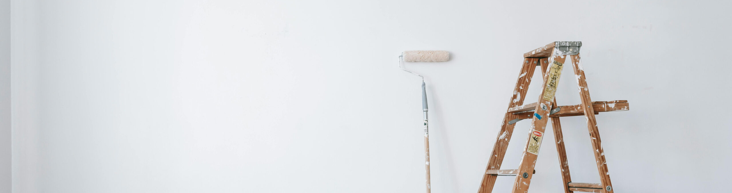 Una pared blanca en renovación muestra un rodillo de pintura apoyado junto a una escalera de madera cubierta de salpicaduras en una estancia luminosa.